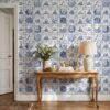 Delft tile wallpaper with bookish medallions behind a carved wooden console table with a linen lamp